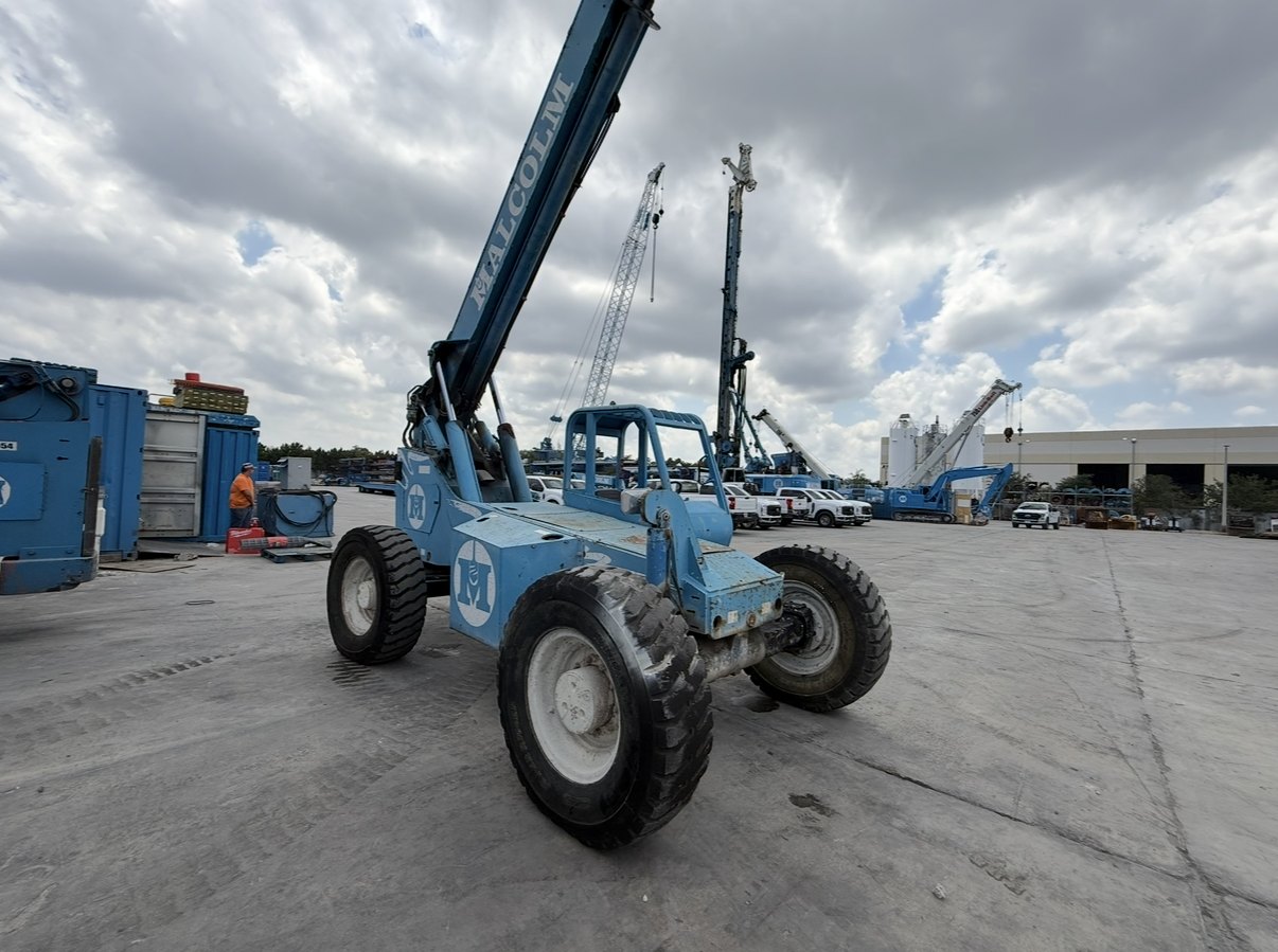 Blue telehandler with extended boom on a contractor yard surrounded by cranes and drill rigs