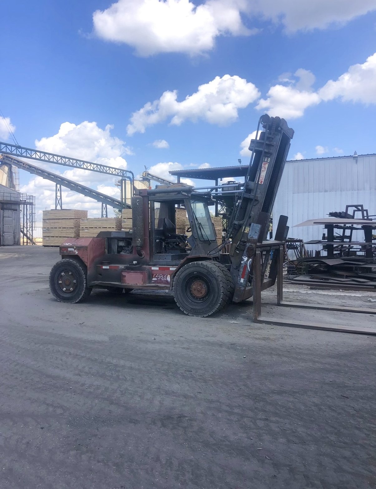 Heavy high-capacity red pneumatic-tire forklift at a lumber mill with conveyor and stacked timber behind